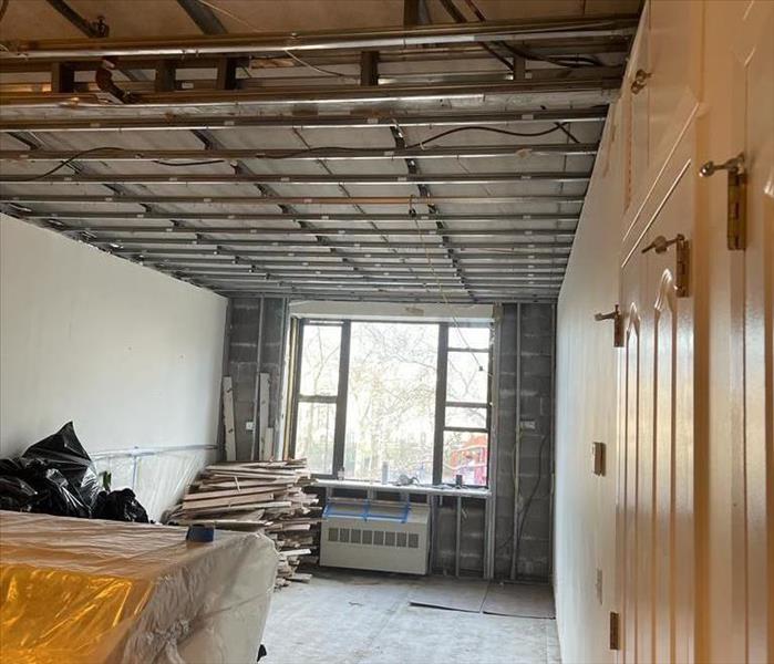 AN apartment with a stripped ceiling and rear wall with radiator and window. Bags of debris and piled-up baseboards from demo