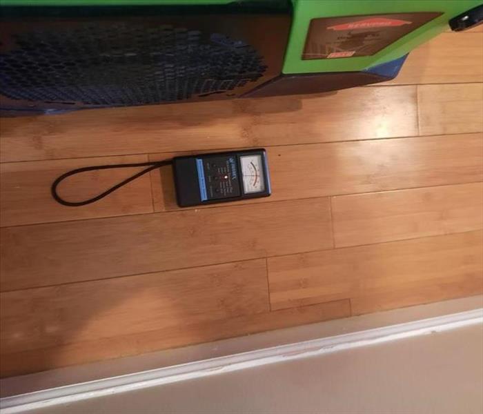 SERVPRO dehumidifier near a moisture meter reading elevated water levels for a wood floor
